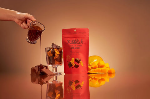 Nibblech mango chocolate product with a hand pouring a drink and a glass of mangoes on a reflective surface.