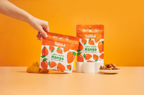 Hand holding a packet of Nibbleish Mango snack with other packets and snacks on an orange background